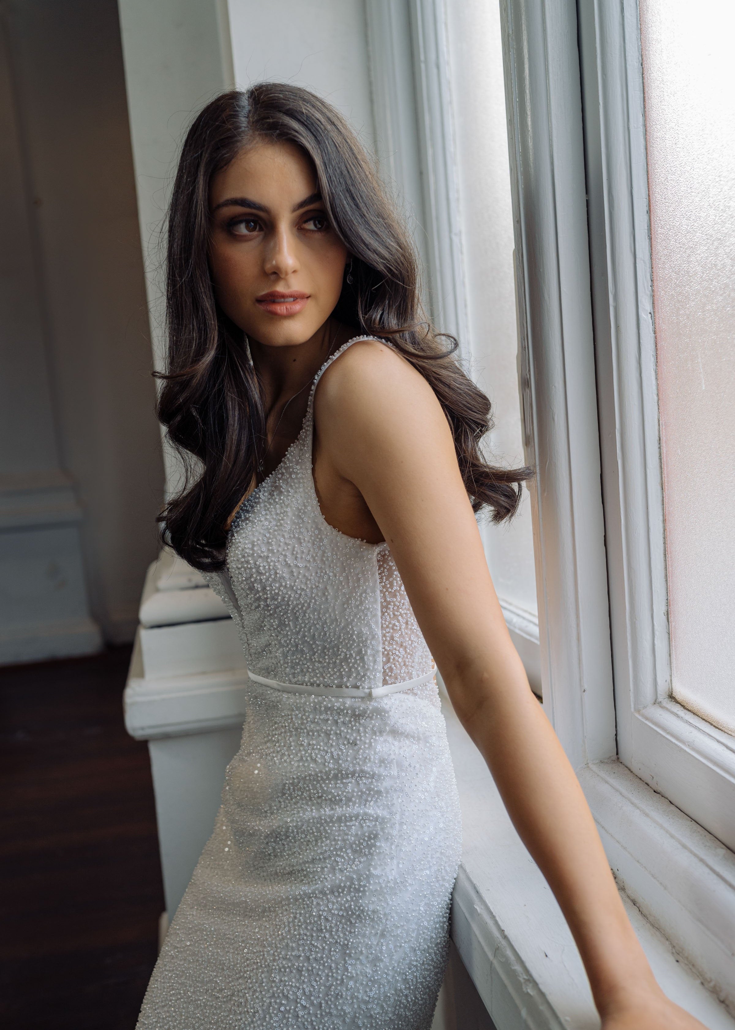 Close-up of bride wearing a beaded fit and flare wedding dress with plunging V neckline standing by a window.