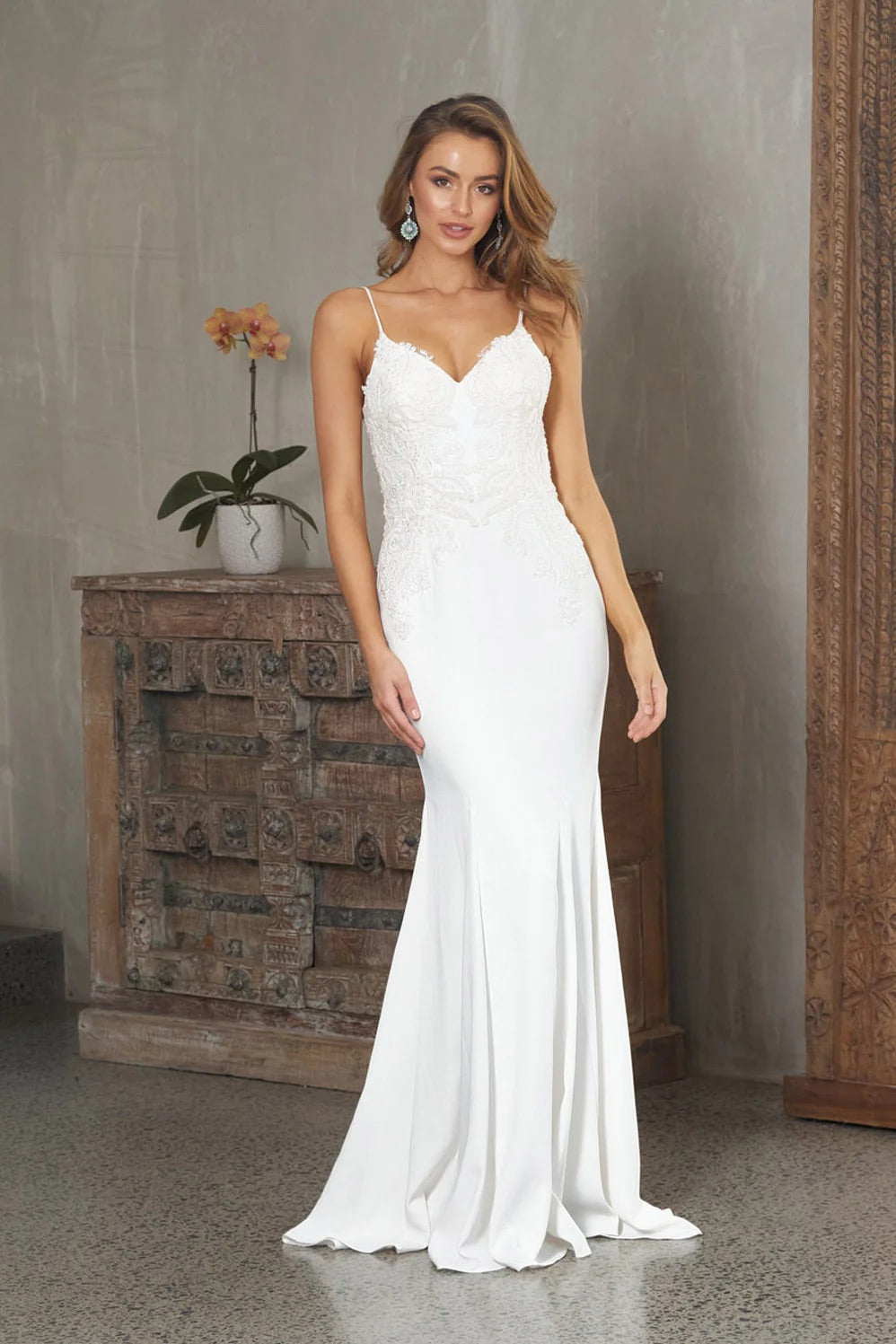 A woman standing in a room wearing a white, V-neck Crepe Fit and Flare Wedding Dress with a plunging back.