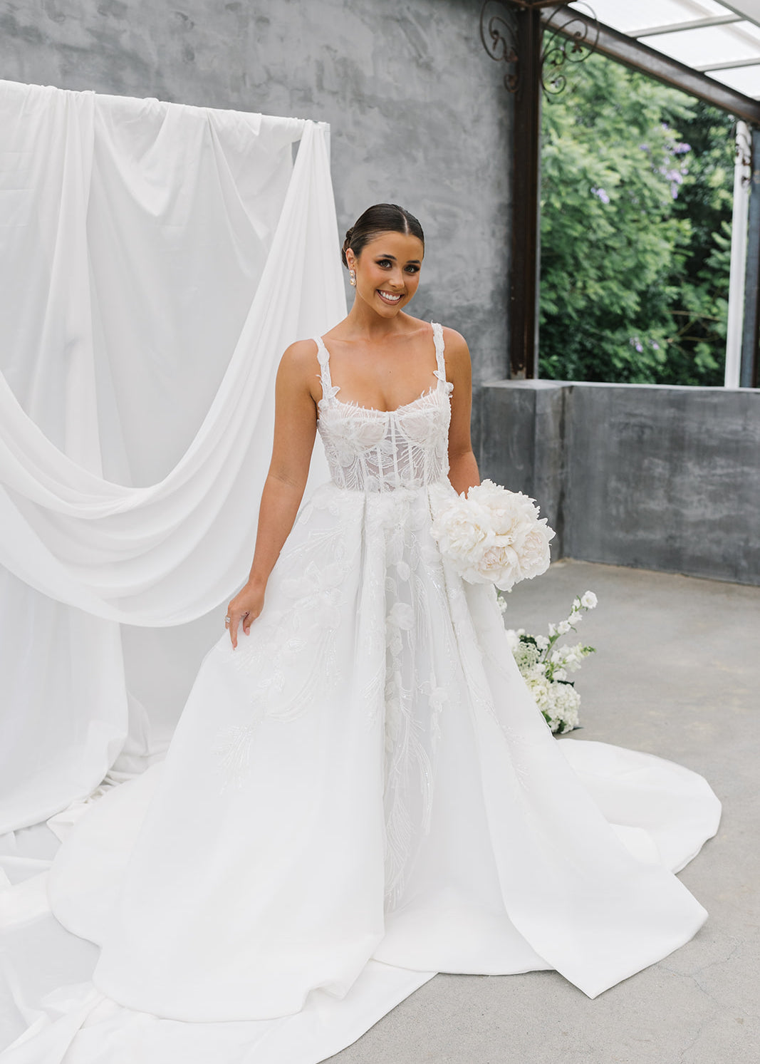 Woman in a A-Line Wedding Dress holding flowers with a white curtain and greenery in the background