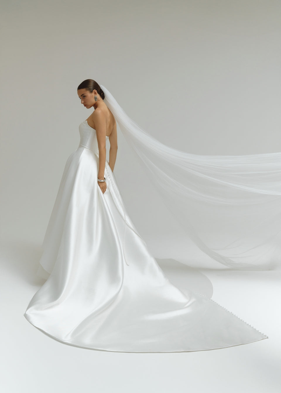 Woman in a strapless Mikado ballgown wedding dress with a long train on a plain background
