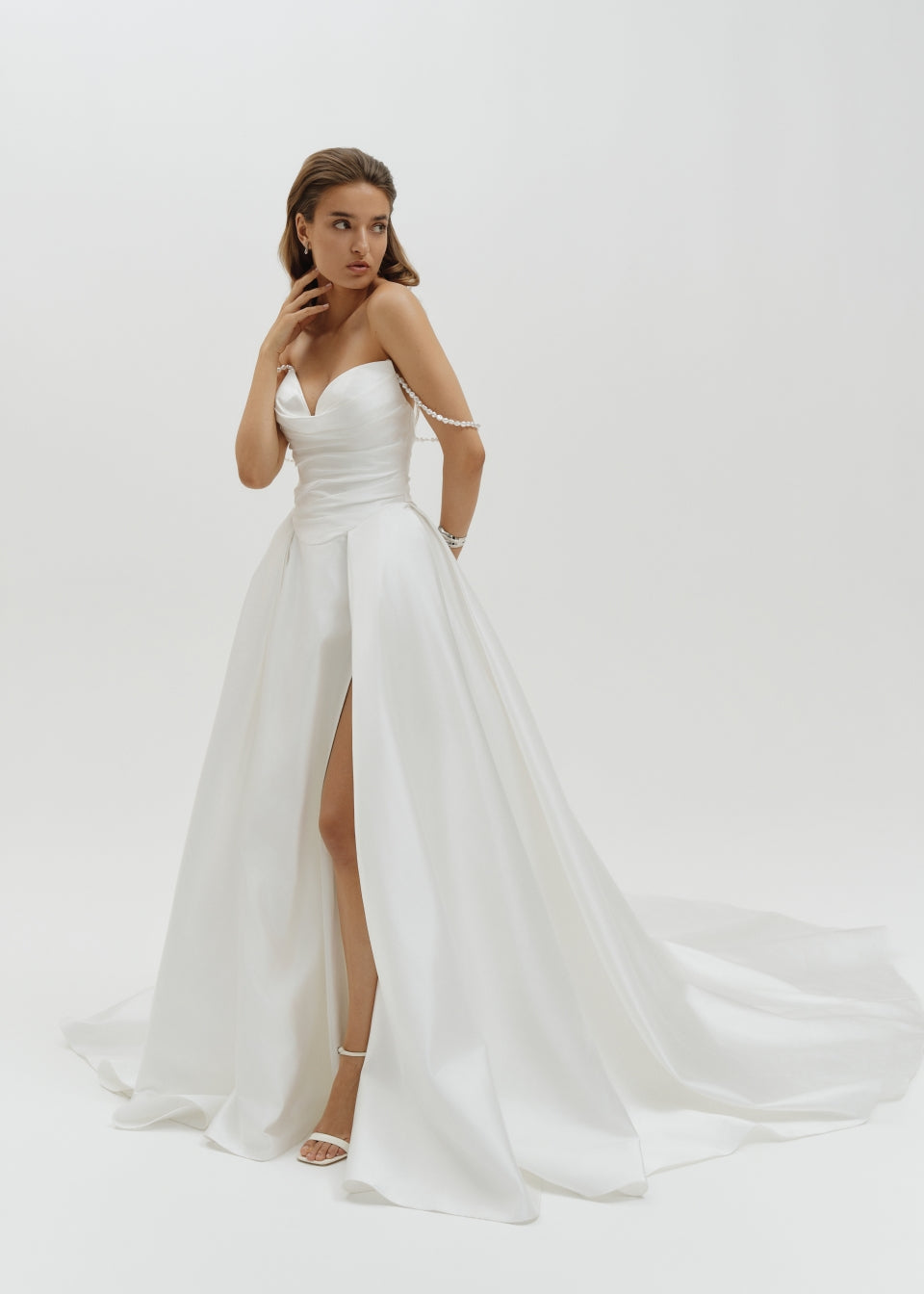 Woman wearing a Duchess satin basque waist wedding dress with a slit on a white background