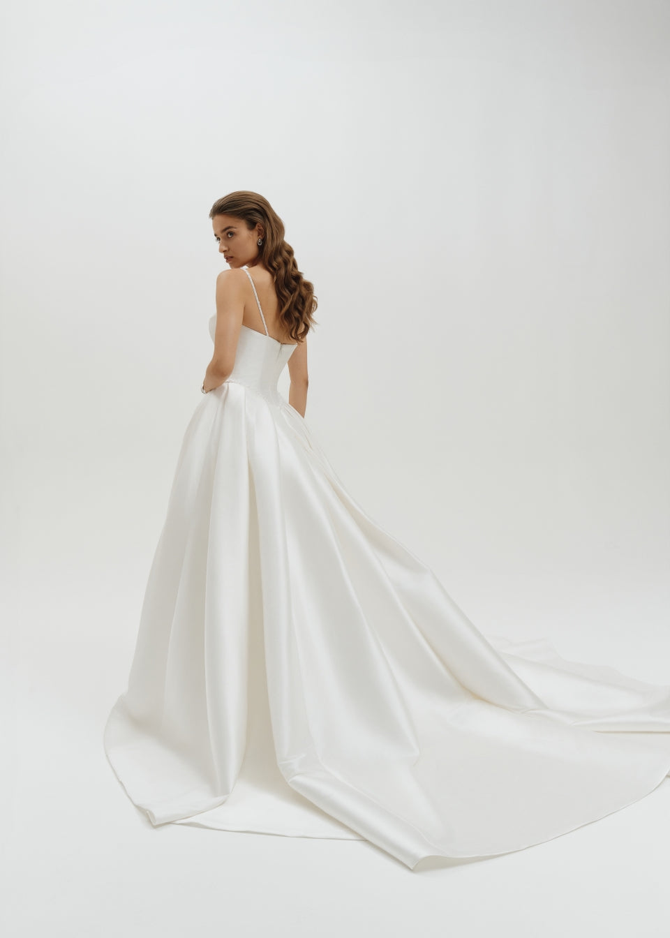 Woman wearing a basque waist Mikado ballgown wedding dress with a long train on a plain background