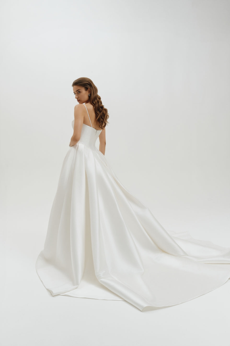 Woman wearing a basque waist Mikado ballgown wedding dress with a long train on a plain background