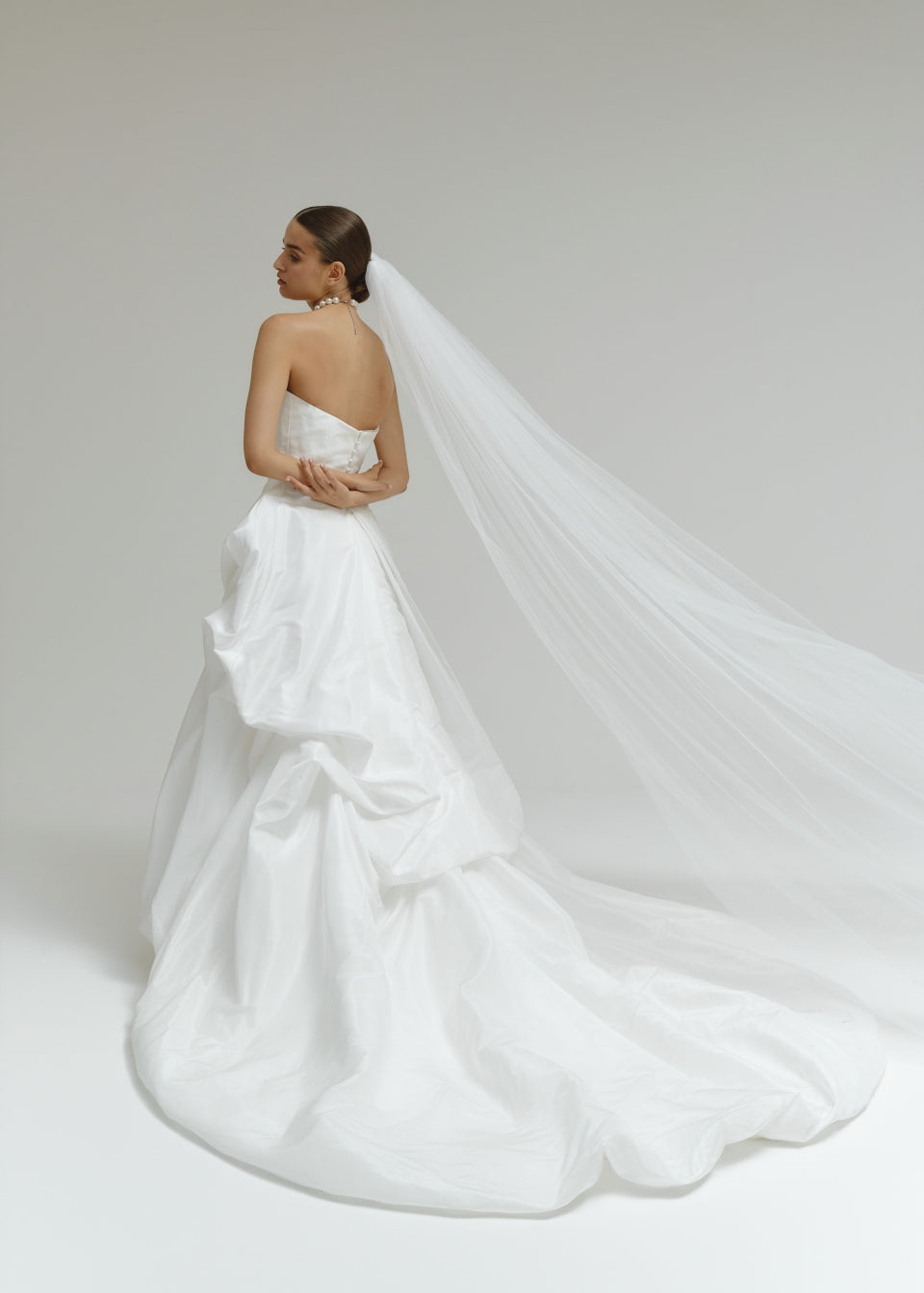 Woman in a strapless matte satin bubble wedding dress with a long veil on a plain background