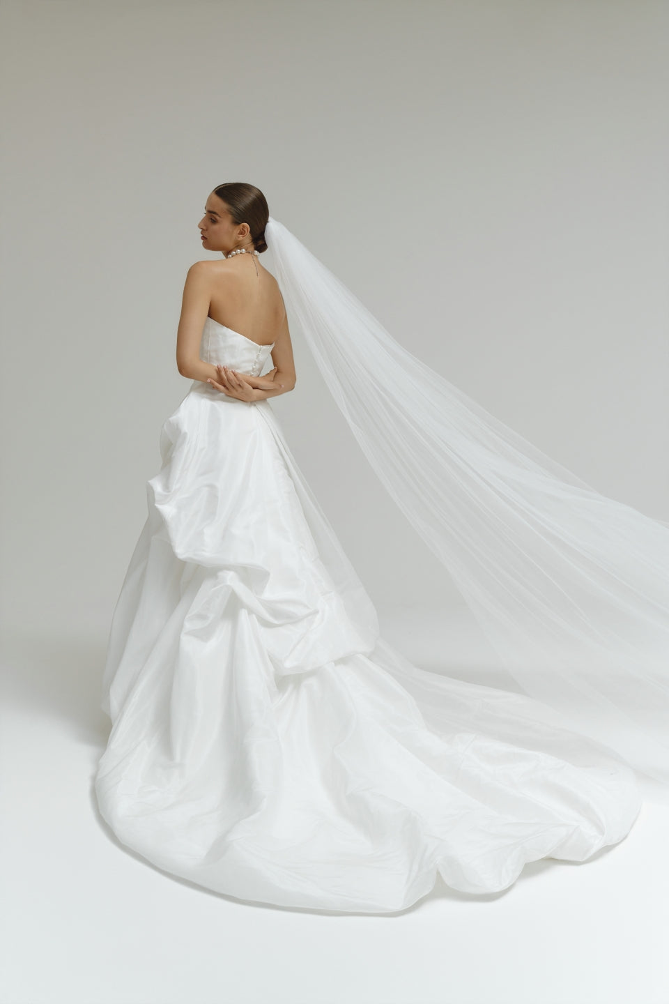 Woman in a strapless matte satin bubble wedding dress with a long veil on a plain background