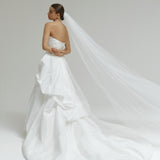 Woman in a strapless matte satin bubble wedding dress with a long veil on a plain background