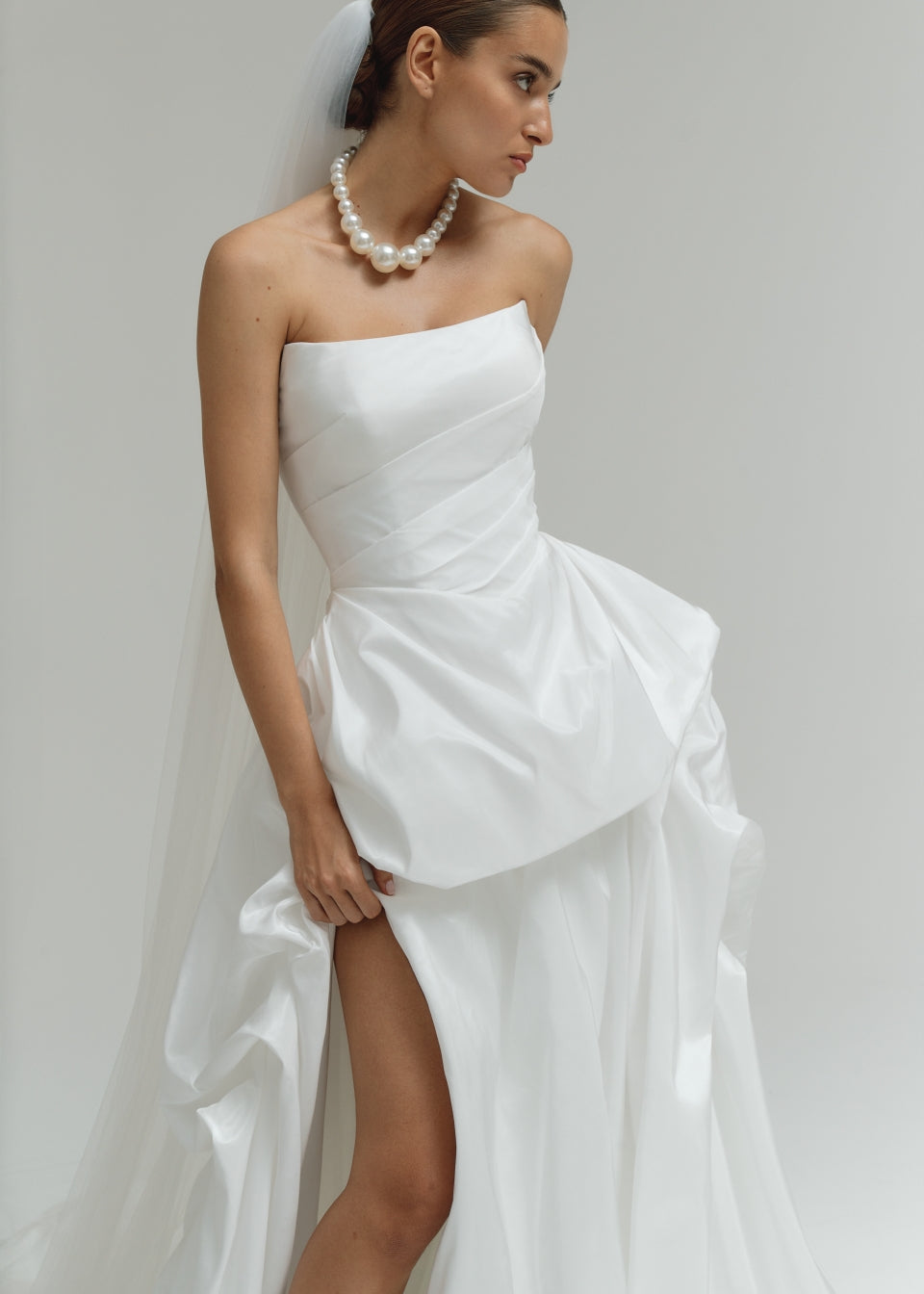 Woman wearing a strapless matte satin bubble wedding dress with a high slit on a plain background