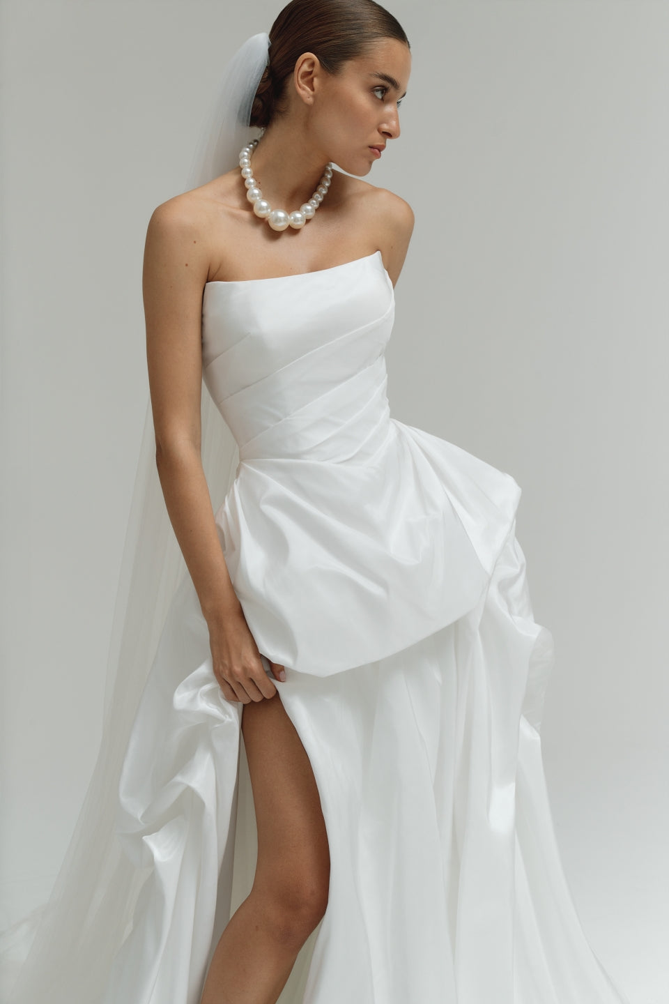 Woman wearing a strapless matte satin bubble wedding dress with a high slit on a plain background