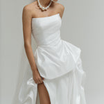 Woman wearing a strapless matte satin bubble wedding dress with a high slit on a plain background