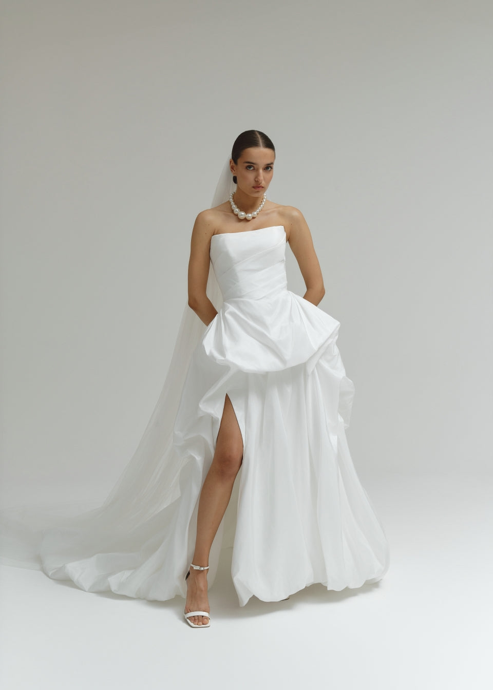 Woman wearing a strapless matte satin bubble wedding dress with a slit on a plain background