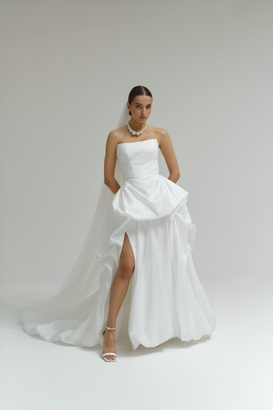 Woman wearing a strapless matte satin bubble wedding dress with a slit on a plain background