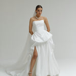 Woman wearing a strapless matte satin bubble wedding dress with a slit on a plain background