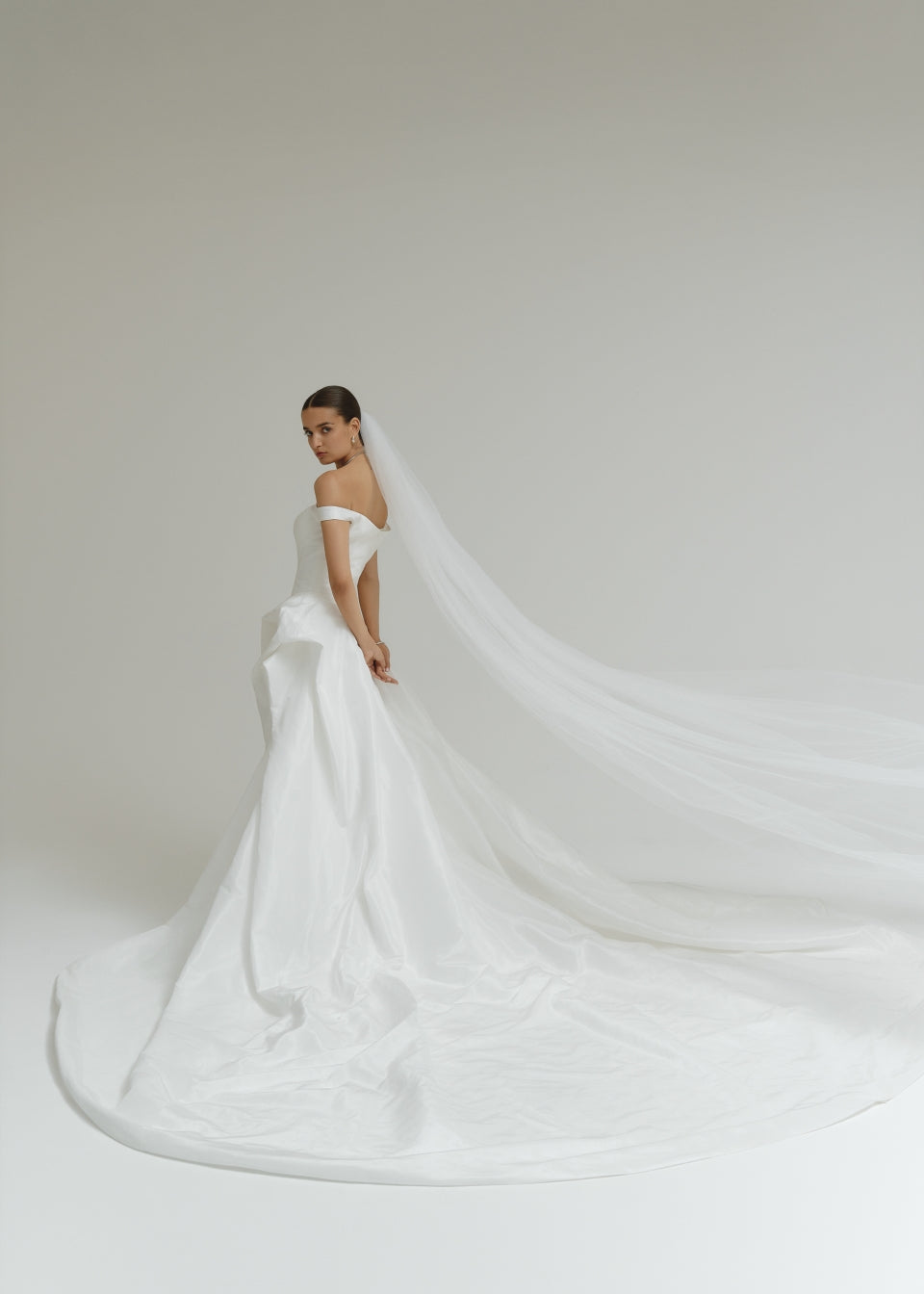 Bride in a off-the-shoulder matte satin ballgown wedding dress with a long veil against a plain background