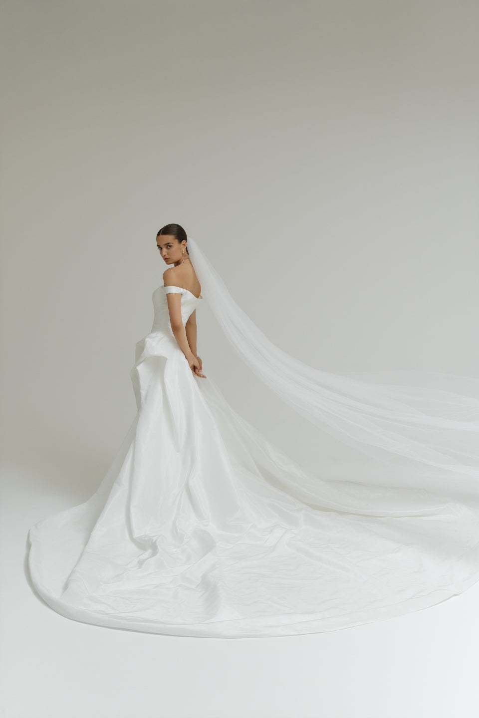 Bride in a off-the-shoulder matte satin ballgown wedding dress with a long veil against a plain background