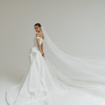 Bride in a off-the-shoulder matte satin ballgown wedding dress with a long veil against a plain background