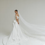 Bride in a off-the-shoulder matte satin ballgown wedding dress with a long veil against a plain background