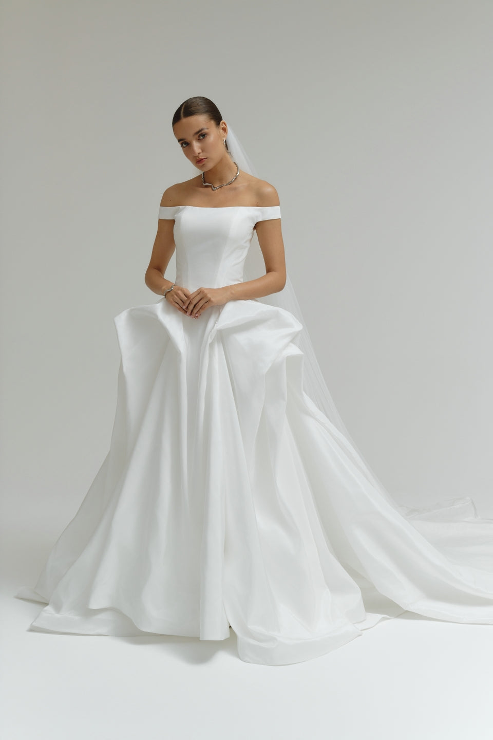 Woman wearing a off-the-shoulder matte satin ballgown wedding dress on a plain background