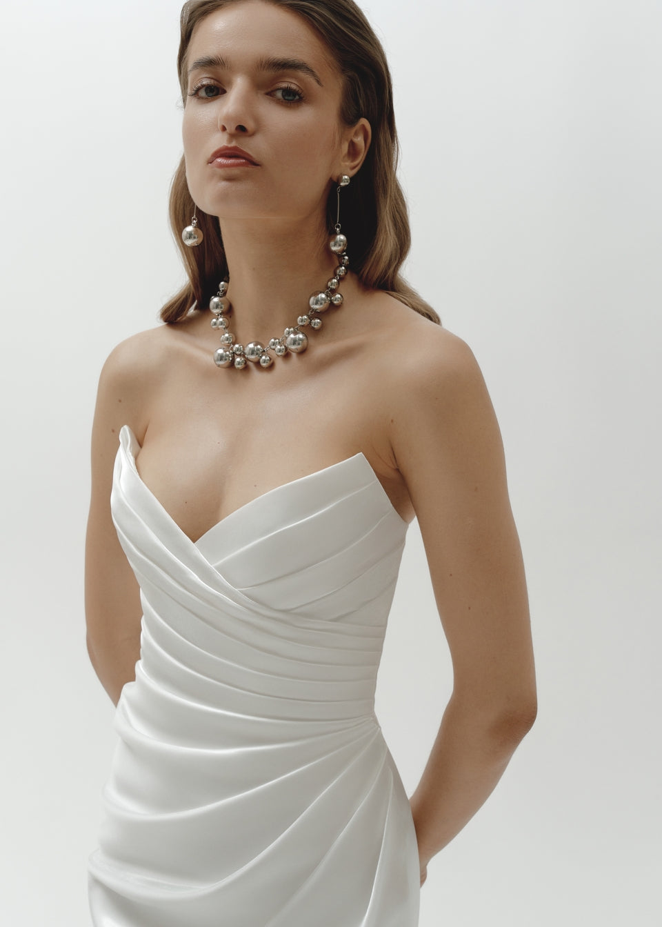 Woman wearing a strapless liquid organza fitted wedding dress and pearl necklace on a plain background