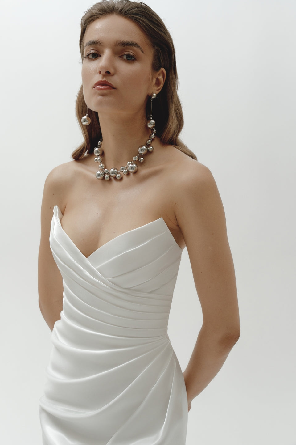 Woman wearing a strapless liquid organza fitted wedding dress and pearl necklace on a plain background