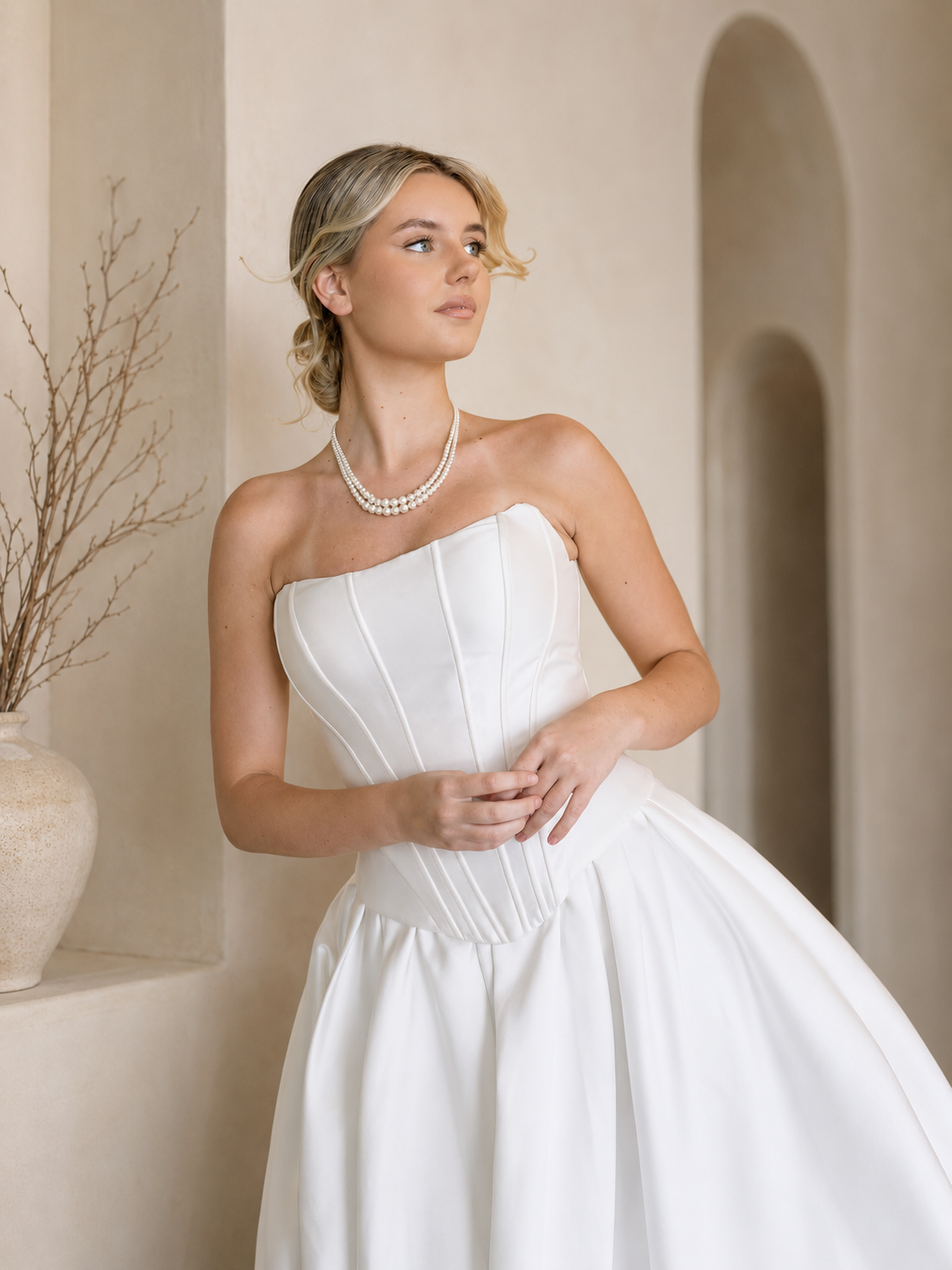 Woman in a Modern Mikado Wedding Dress with a pearl necklace against a warm-toned wall.
