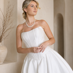 Woman in a Modern Mikado Wedding Dress with a pearl necklace against a warm-toned wall.