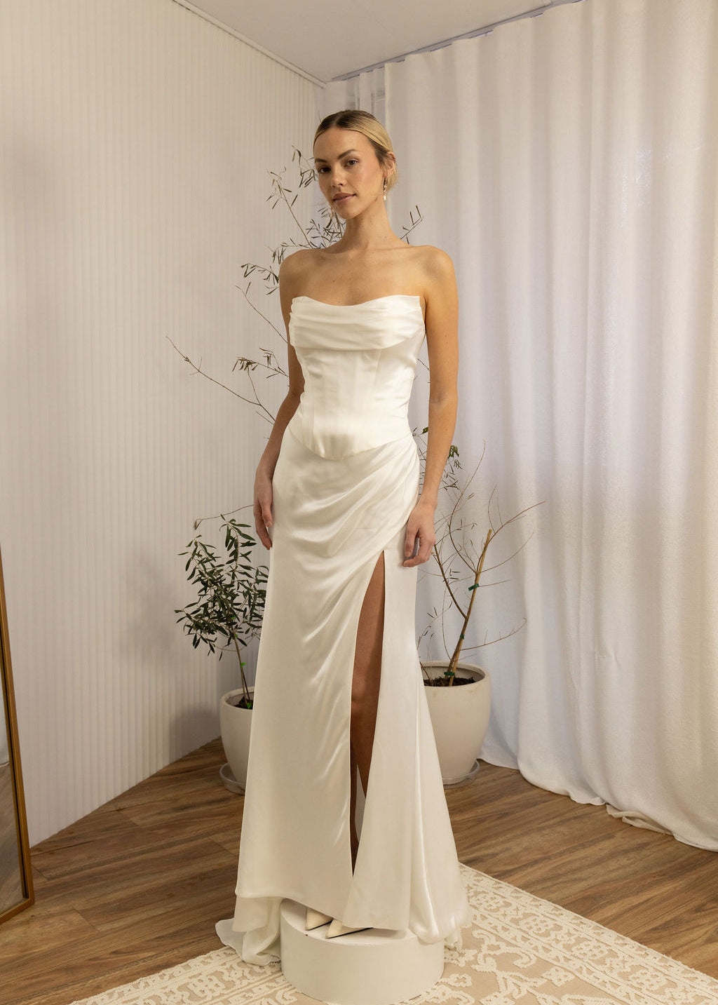 Woman wearing a strapless Two Piece Bridal Gown with a high slit in a room with curtains and plants.