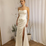 Woman wearing a strapless Two Piece Bridal Gown with a high slit in a room with curtains and plants.