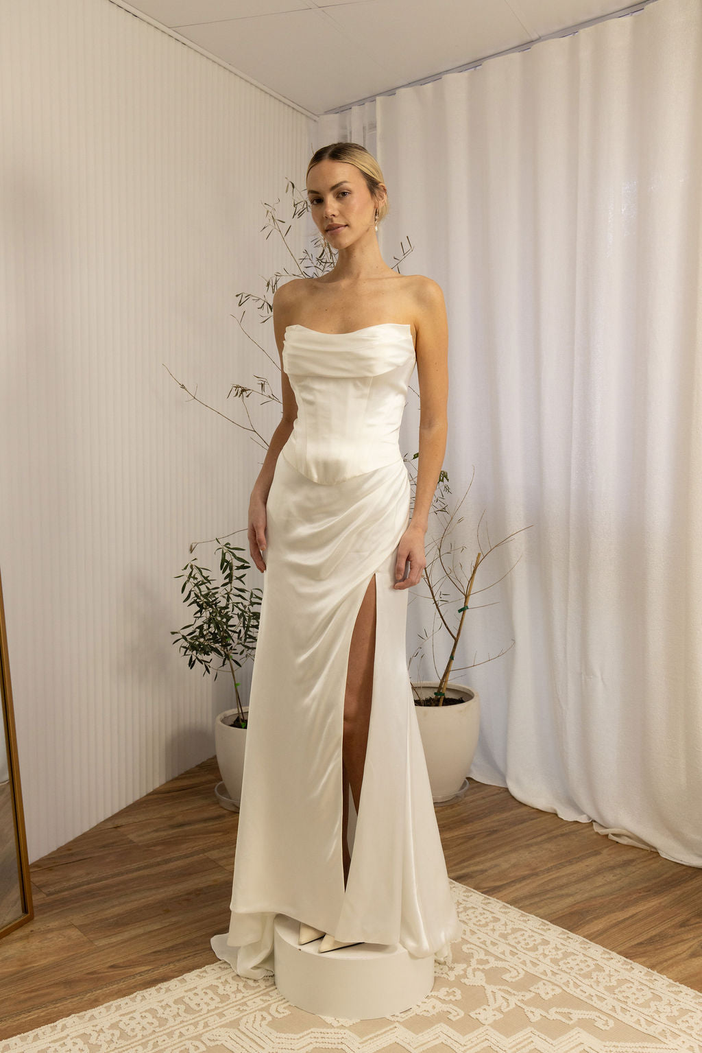 Woman wearing a strapless Two Piece Bridal Gown with a high slit in a room with curtains and plants.