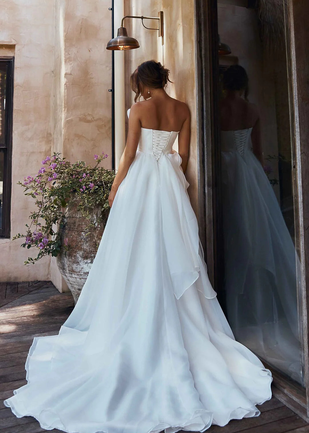 Woman in an organza A-line wedding dress standing in a sunlit room with flowers and a mirror.