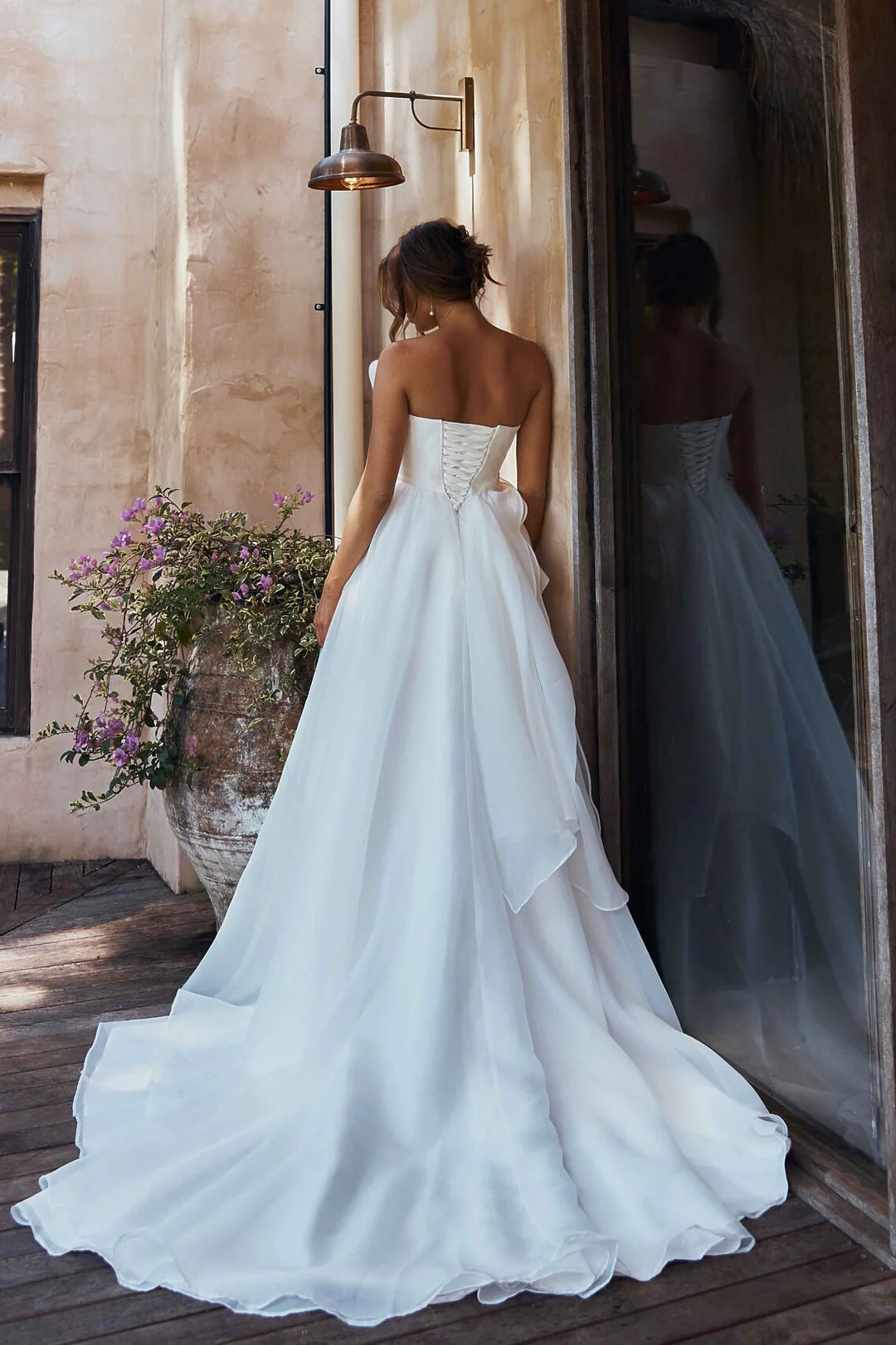 Woman in an organza A-line wedding dress standing in a sunlit room with flowers and a mirror.