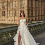 Woman in an organza A-line wedding dress with a high slit standing on a bridge with a large stone building in the background.