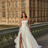Woman in an organza A-line wedding dress with a high slit standing on a bridge with a large stone building in the background.