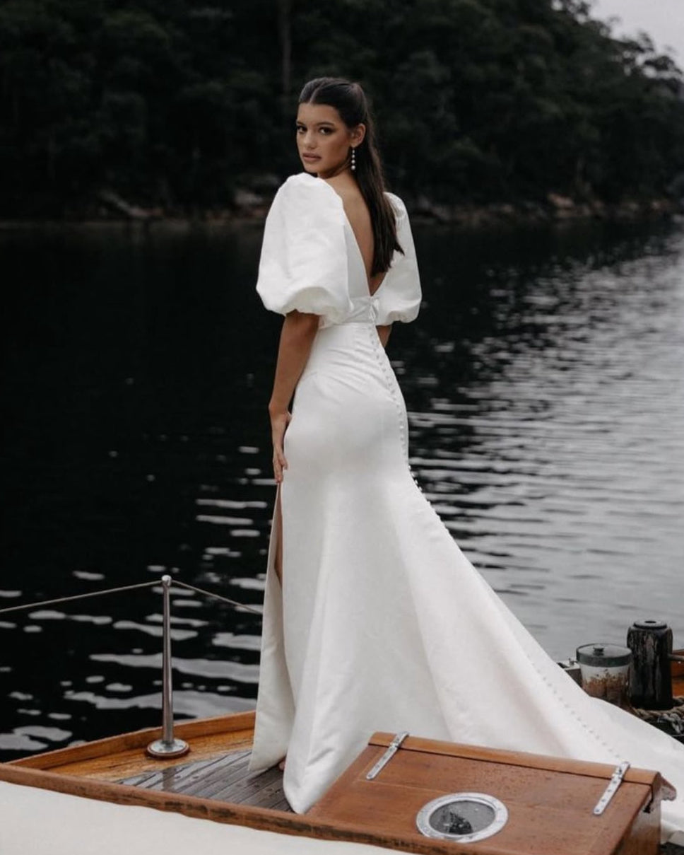 Bride wearing a satin fit and flare wedding dress with puff sleeves and deep V-back standing on a boat by the water.