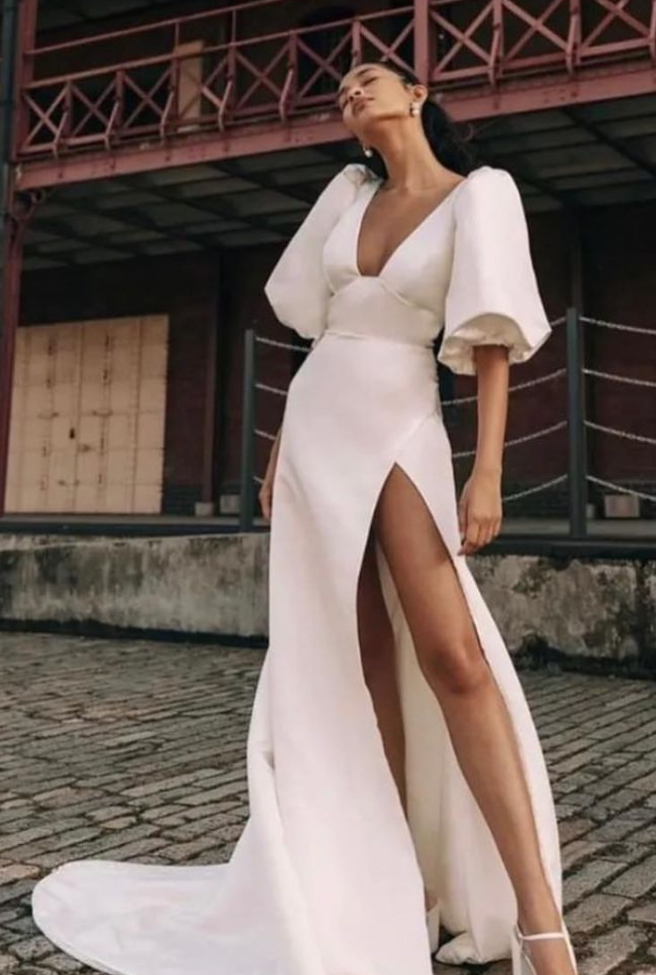 Bride wearing a satin wedding dress with puff sleeves, deep V neckline, and high leg slit standing in an urban setting.