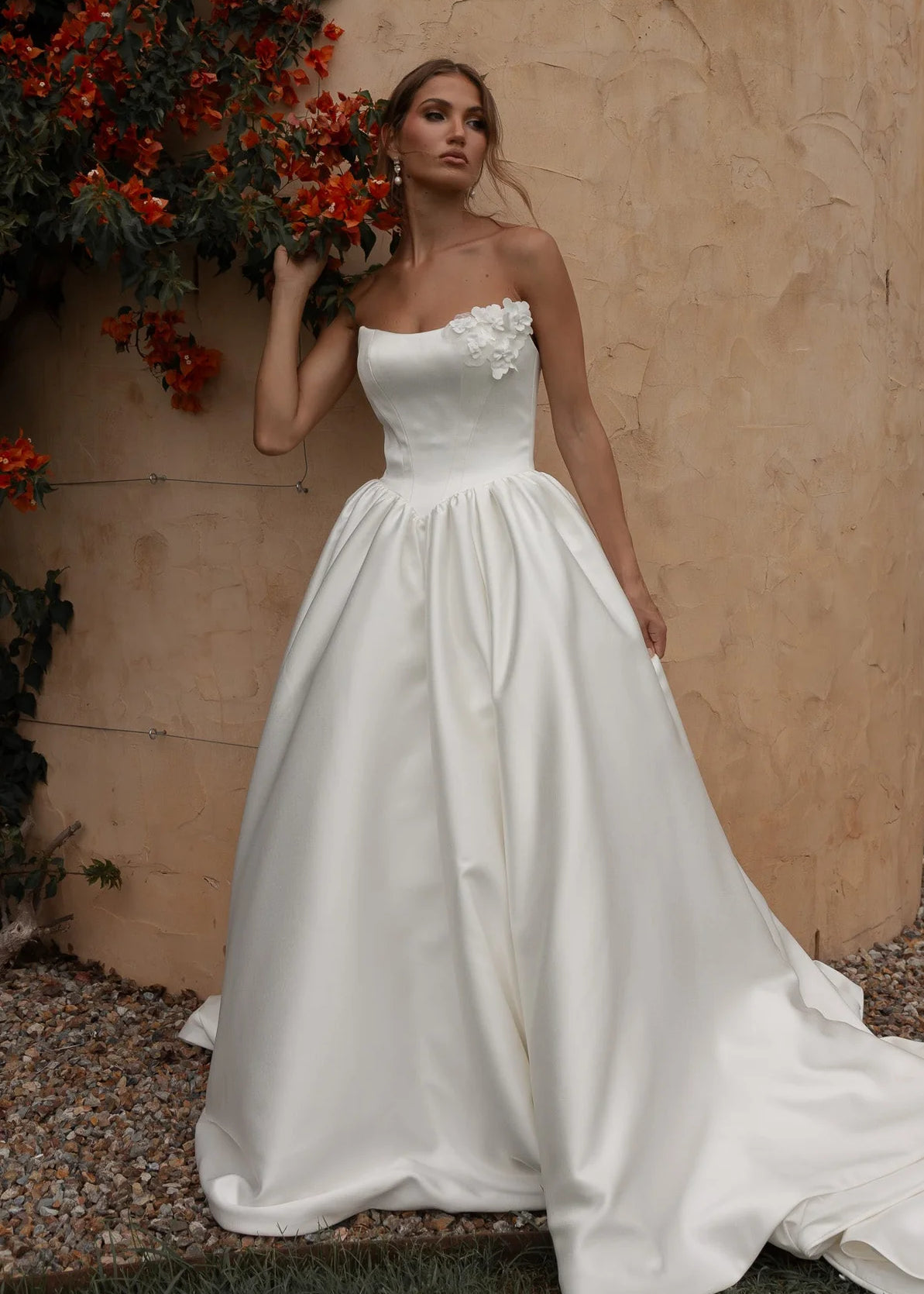 Woman in a Mikado A-Line Wedding Dress standing against a textured wall with red flowers.