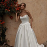 Woman in a Mikado A-Line Wedding Dress standing against a beige wall with red flowers.