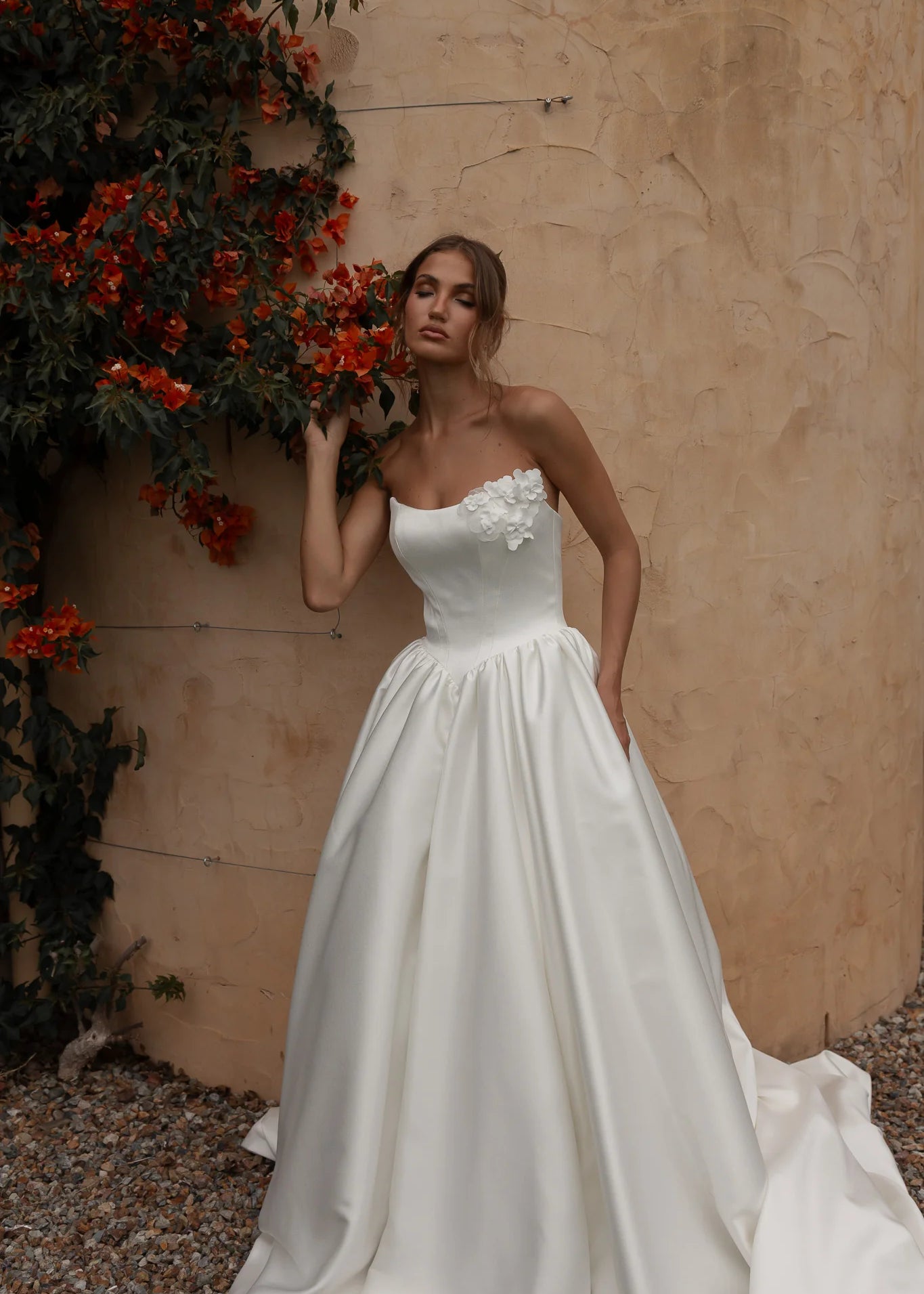 Woman in a Mikado A-Line Wedding Dress standing against a beige wall with red flowers.