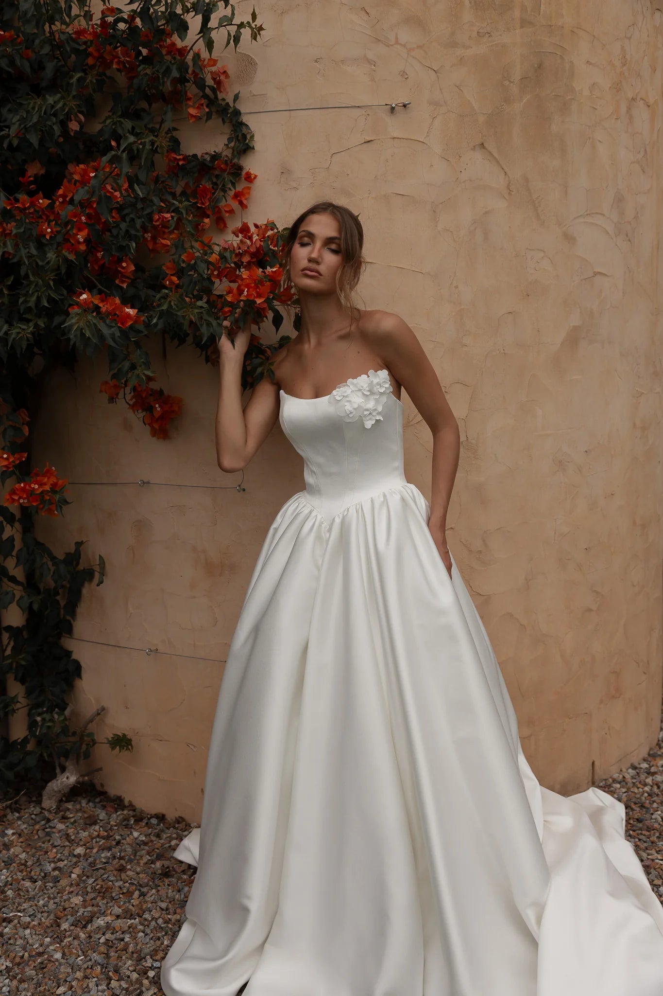 Woman in a Mikado A-Line Wedding Dress standing against a beige wall with red flowers.