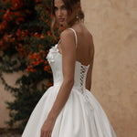 Woman in a Mikado A-Line Wedding Dress with a lace-up back, standing against a beige wall and greenery.
