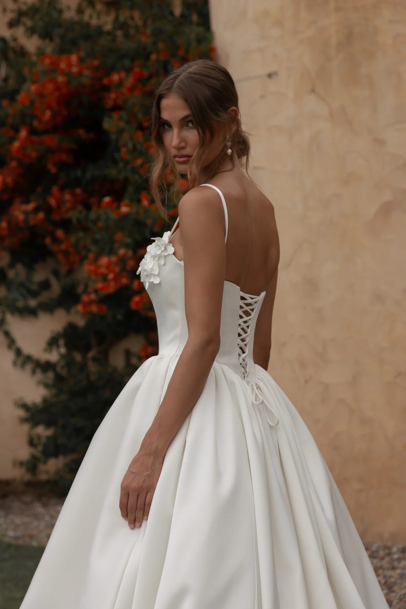 Woman in a Mikado A-Line Wedding Dress with a lace-up back, standing against a beige wall and greenery.