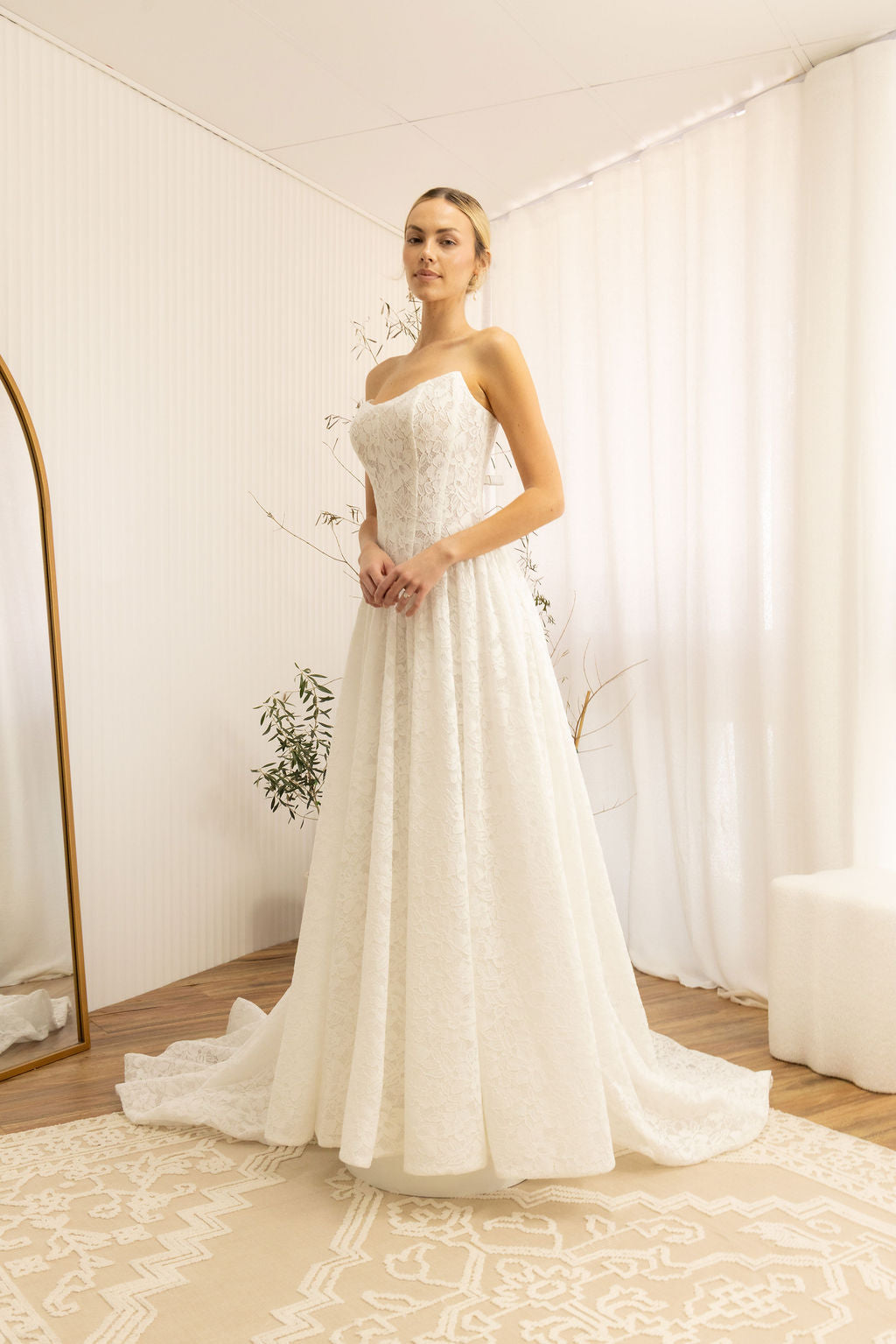 Woman wearing a white Lace Strapless A-Line Wedding Dress in a room with white walls and curtains.
