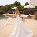 Woman in Genesis wedding dress by Modeca standing by a pool with greenery and umbrellas in the background