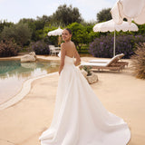 Woman in Genesis wedding dress by Modeca standing by a pool with greenery and umbrellas in the background