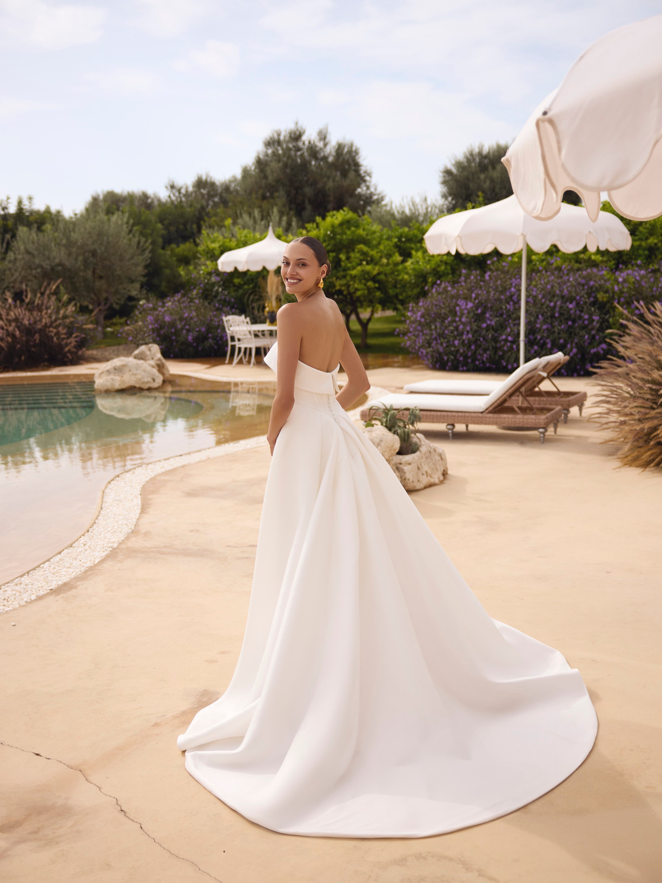 Woman in Genesis wedding dress by Modeca standing by a pool with greenery and umbrellas in the background