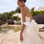 Woman in Genesis wedding dress by Modeca with a large bow standing by a poolside.
