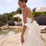 Woman in Genesis wedding dress by Modeca with a large bow standing by a poolside.