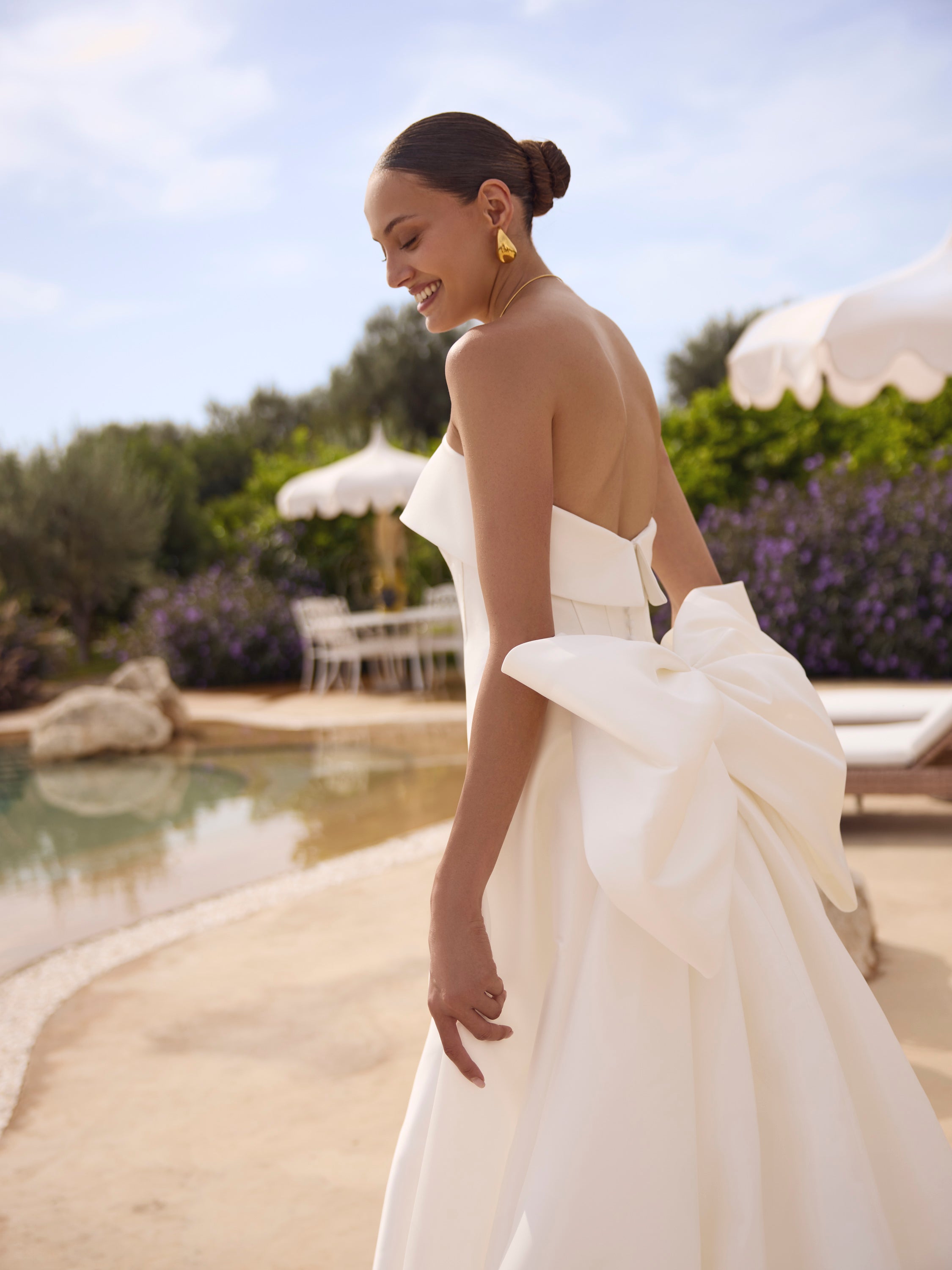 Woman in Genesis wedding dress by Modeca with a large bow standing by a poolside.