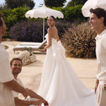 Woman in Genesis wedding dress by Modeca standing outdoors with two men, surrounded by white umbrellas and greenery.