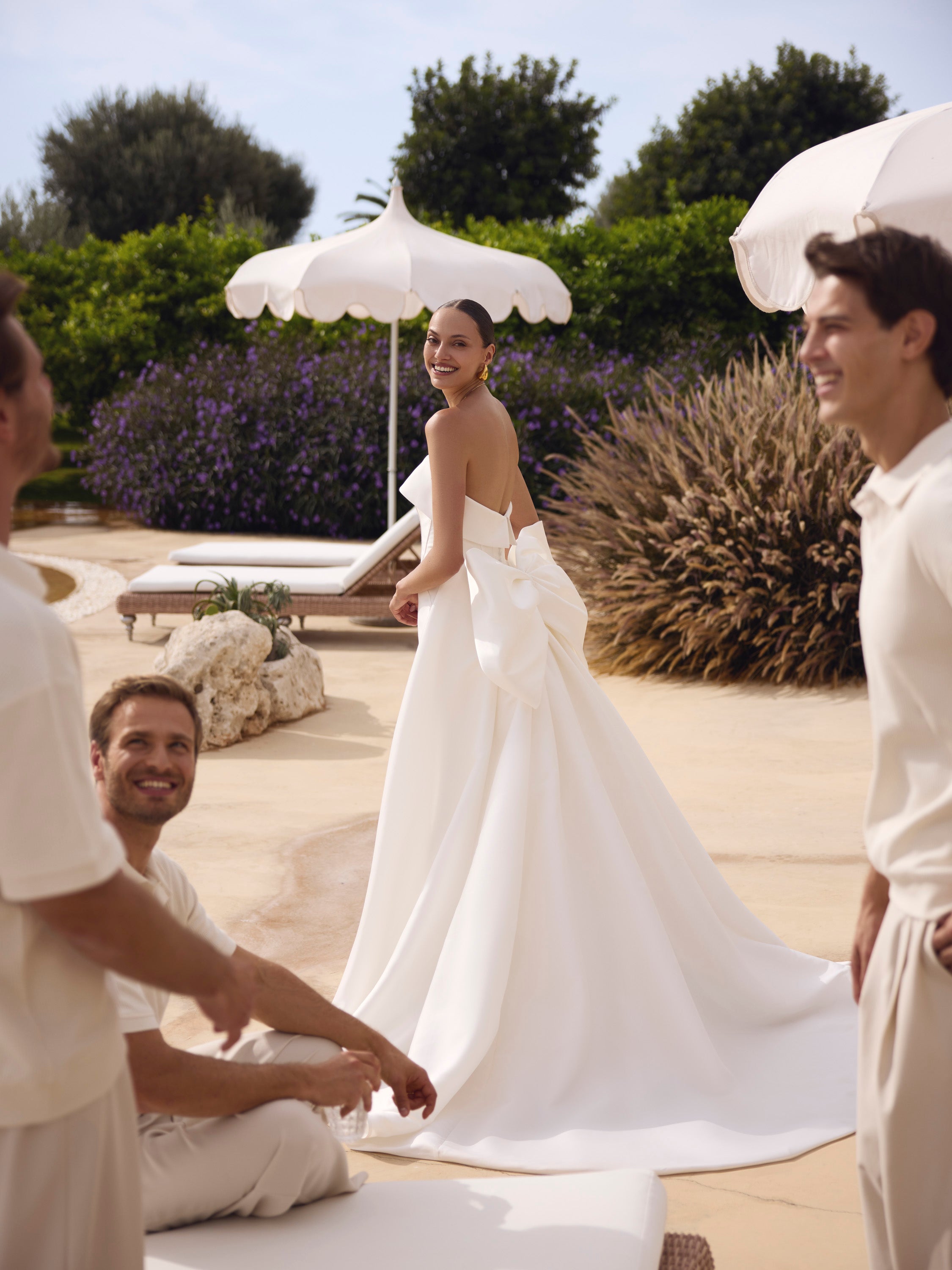 Woman in Genesis wedding dress by Modeca standing outdoors with two men, surrounded by white umbrellas and greenery.