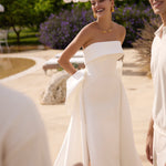 Woman in Genesis wedding dress by Modeca standing outdoors with greenery and a white umbrella in the background.